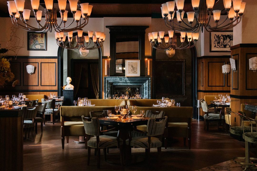 A dimly lit dining room with velvet booths, a marble fireplace, and large glass chandeliers.