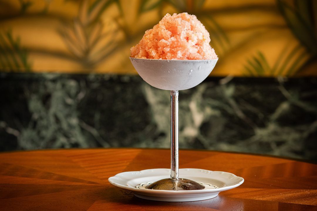 A shaved ice dessert in a frosted glass on a stemmed base, set on a table.
