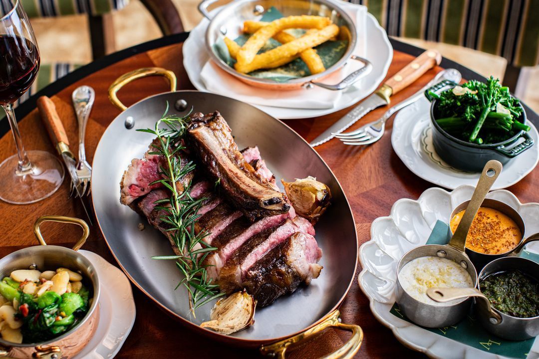 A platter of sliced grilled meat with rosemary, served alongside fries, greens, and a glass of red wine.