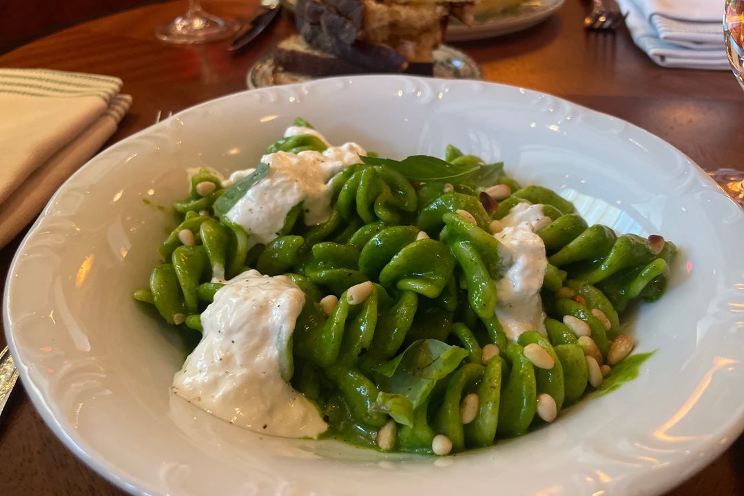 A bowl of green pasta with dollops of burrata and pine nuts.