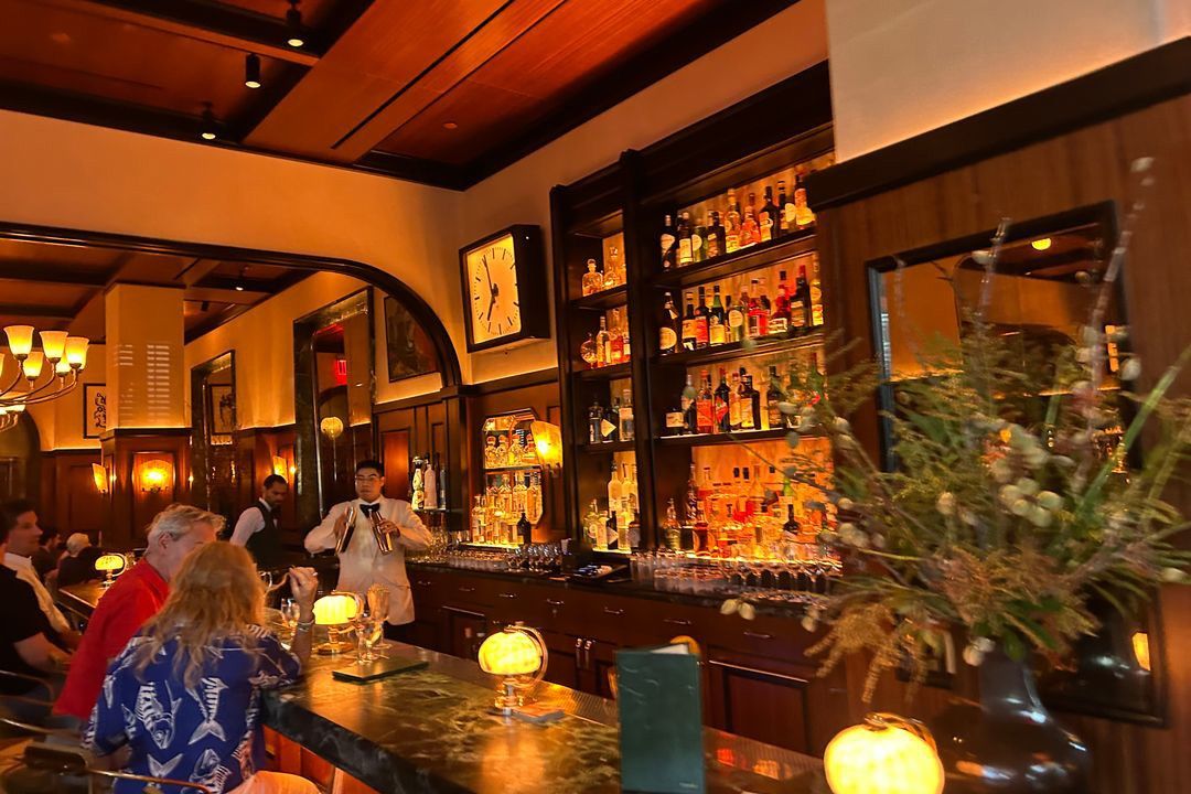 A bartender preparing a drink at a well-stocked bar with diners seated nearby.