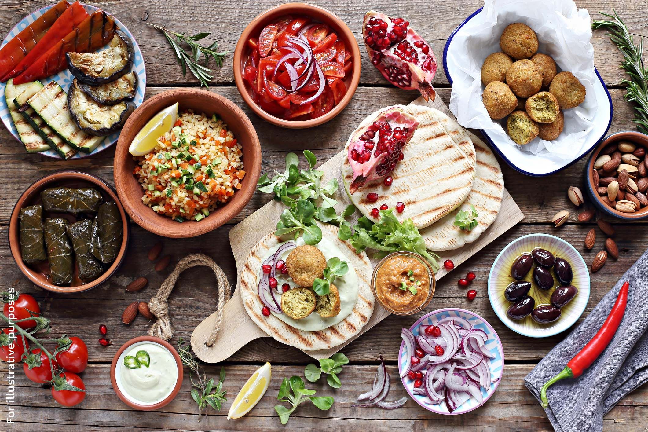 A spread of Mediterranean dishes including grilled vegetables, stuffed vine leaves, falafel, pomegranate, tomato salad, and pitta bread.