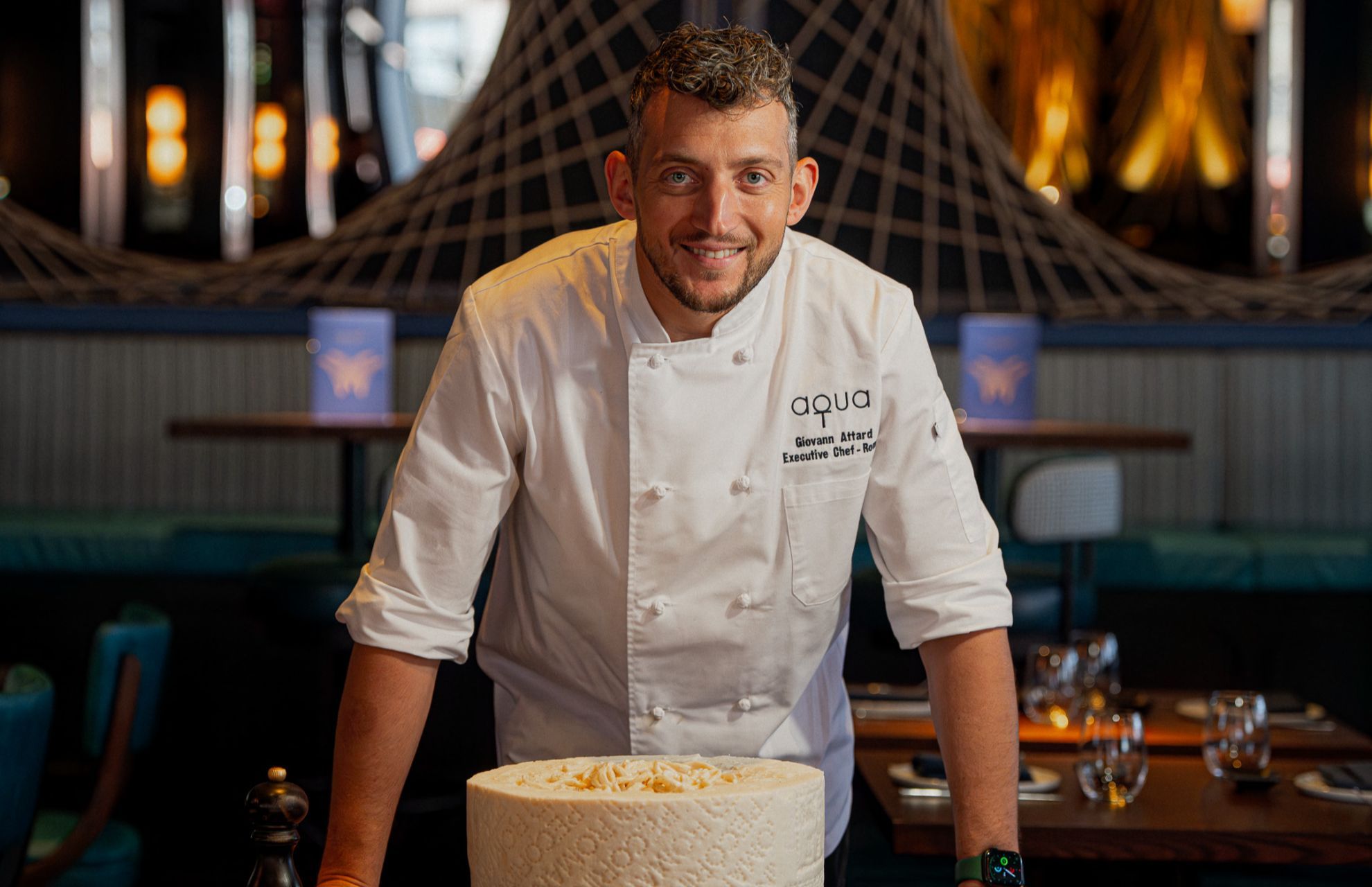 A chef in a white jacket standing behind a wheel of cheese topped with pasta, smiling in a dimly lit dining room.