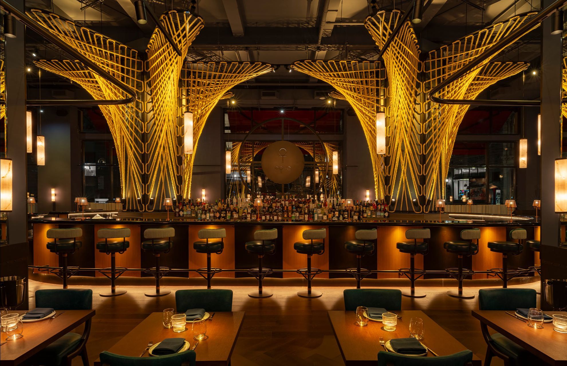 A dining room with a curved bar backed by tall woven gold light fixtures, bottles lined along the counter and set tables in the foreground.