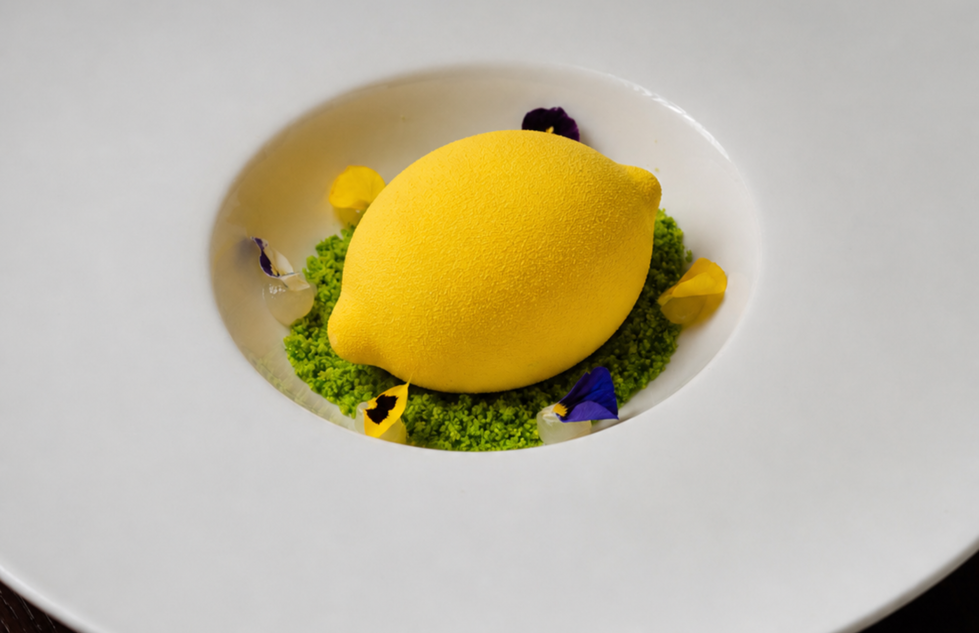 A lemon-shaped dessert with a textured yellow coating, set on a bed of green crumble and garnished with edible flowers.