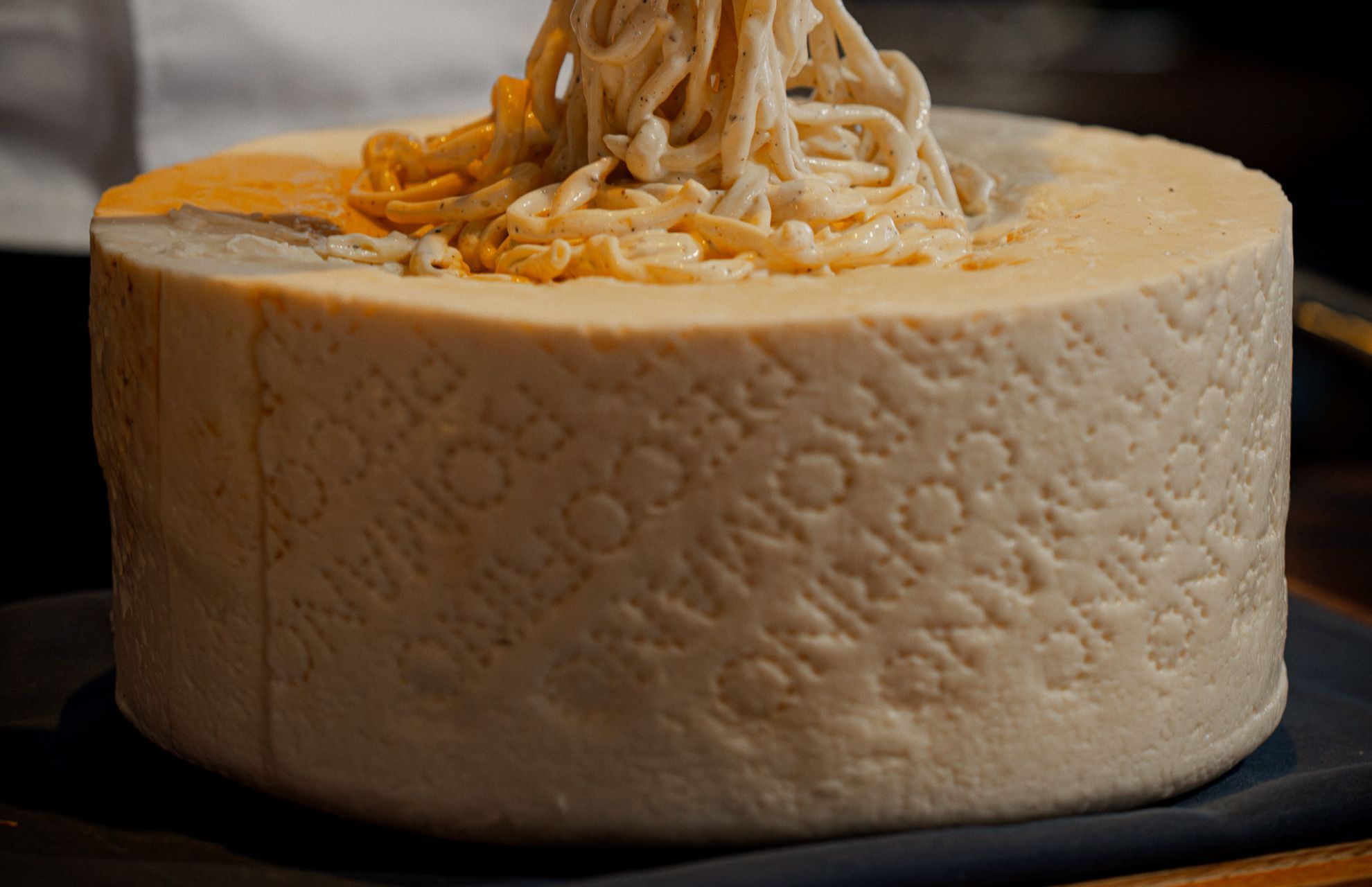 A close-up of a whole wheel of cheese with a patterned rind, topped with pasta being pulled from its centre.