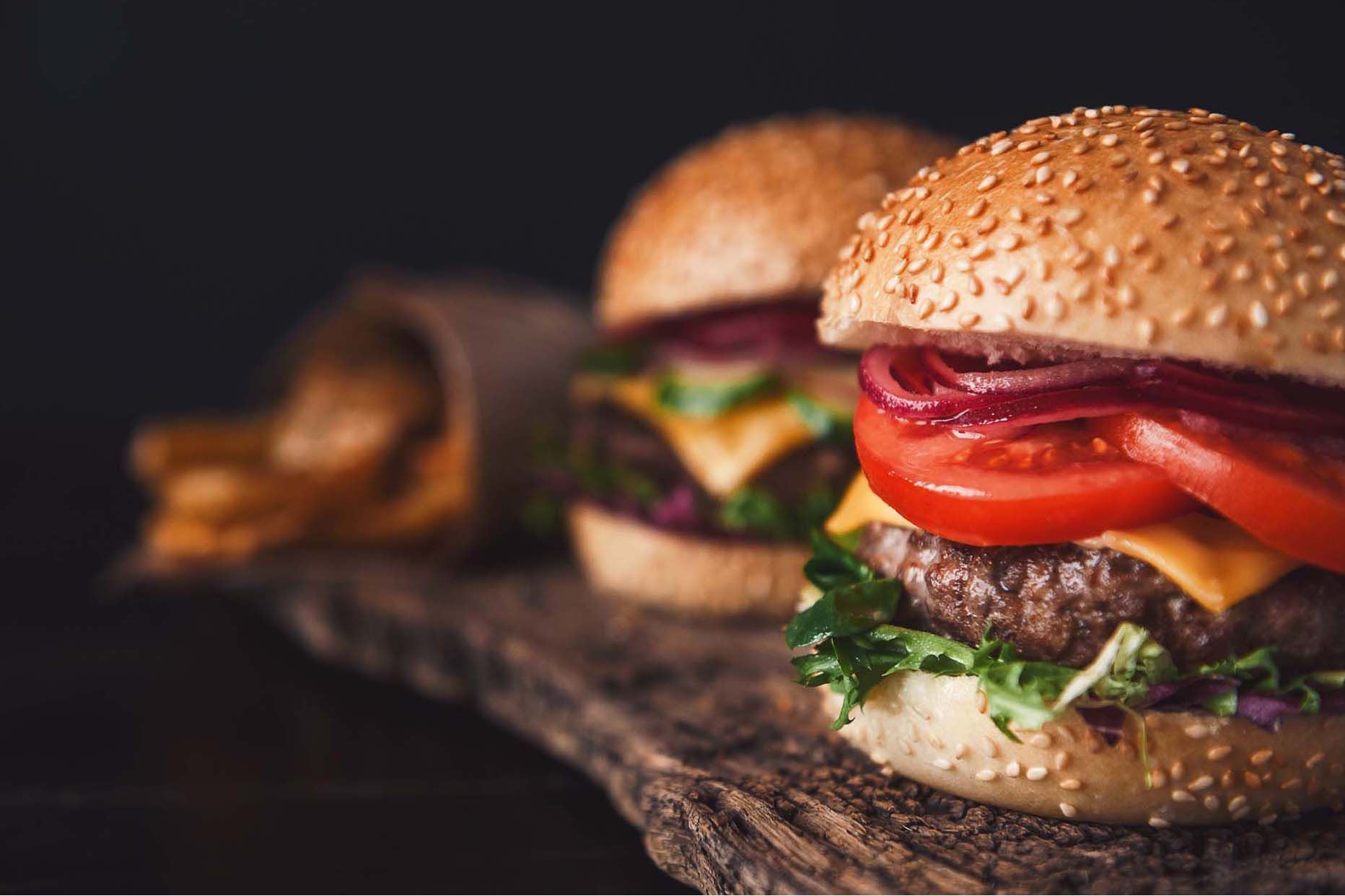 Two burgers with beef patties, melted cheese, tomato, and pickled onion on sesame buns, on a wooden board.