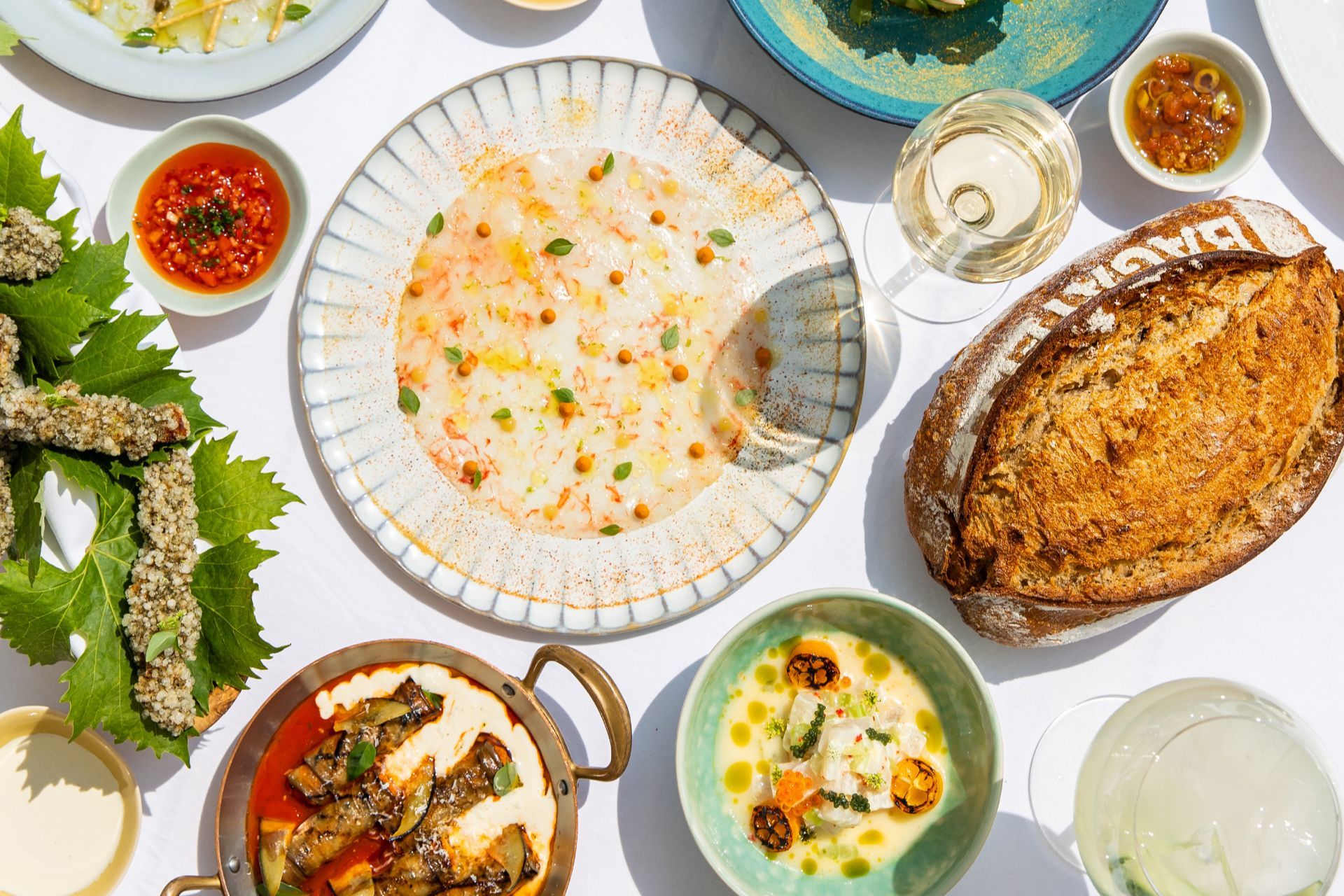 An overhead view of a table spread with seafood dishes, a stuffed vegetable dish, dips, bread and a glass of white wine.