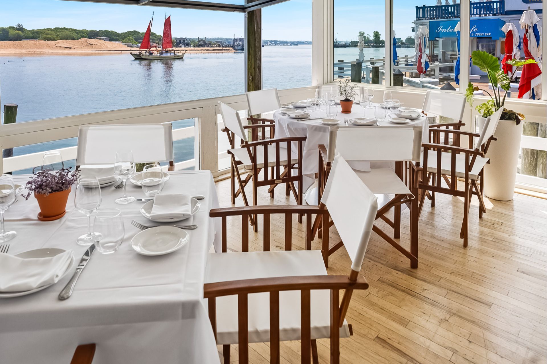 A waterfront dining area with white tablecloths and director's chairs, overlooking a harbor with a red-sailed boat.