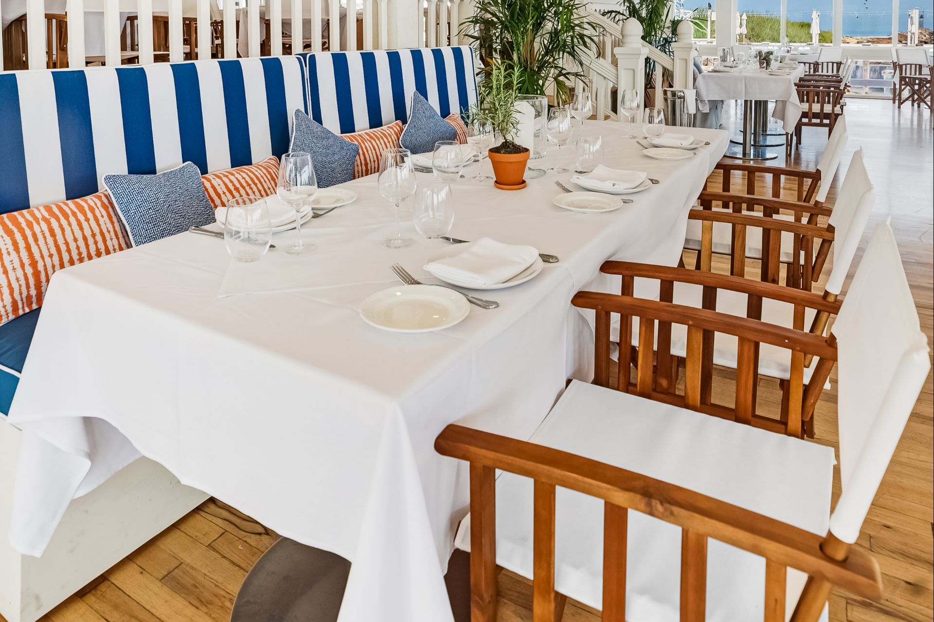 A long table set with white linens, striped and patterned cushions on a bench, and a potted plant, in a bright dining room.
