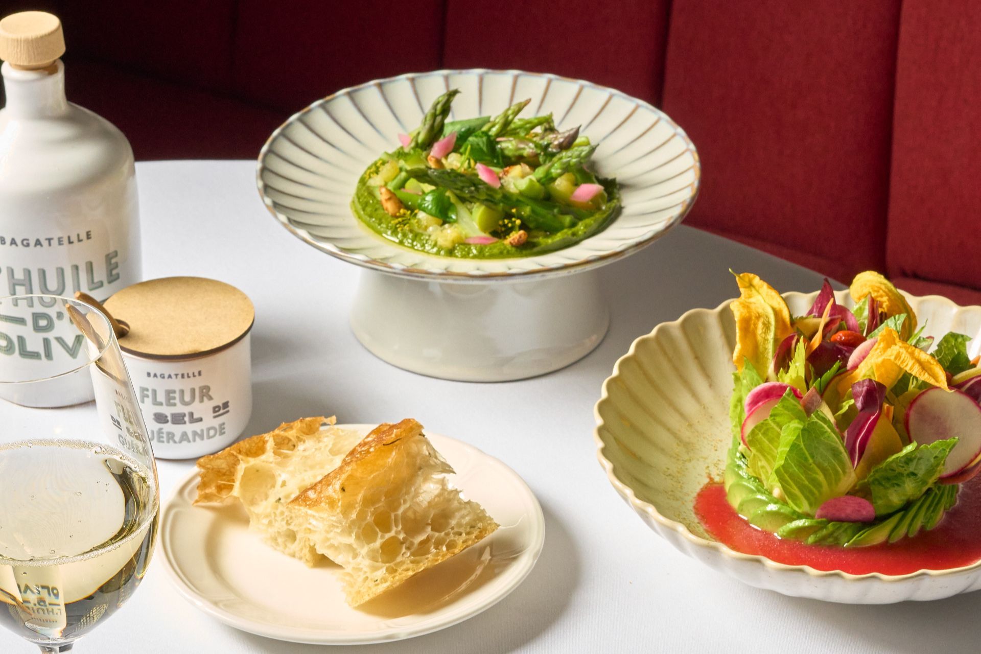 A table with a green vegetable dish on a pedestal plate, a salad with radish and edible flowers, bread, olive oil and seasoning jars, and glasses of wine.