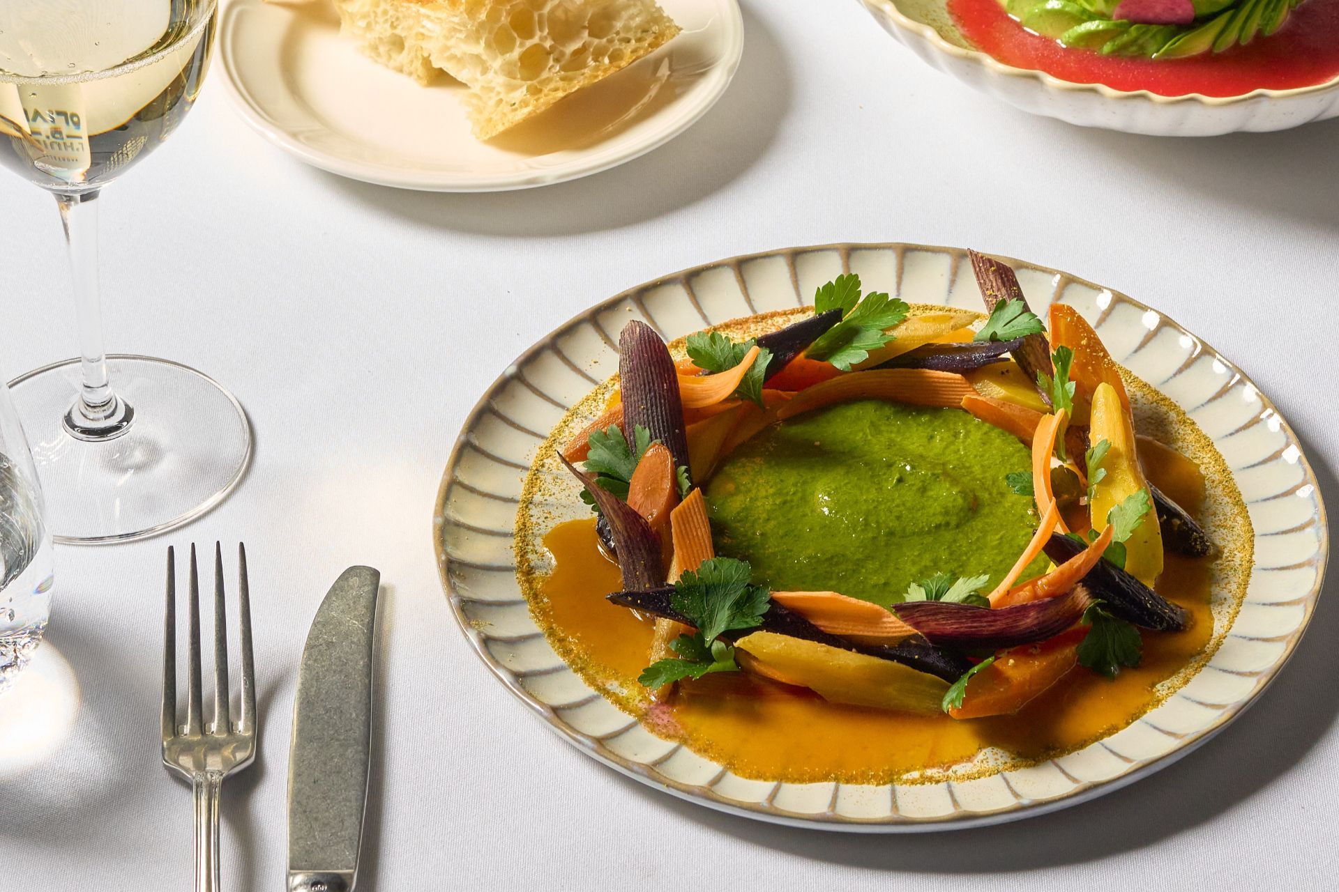 A plated dish of roasted carrots and herbs arranged around a green sauce, with bread and wine on the table.
