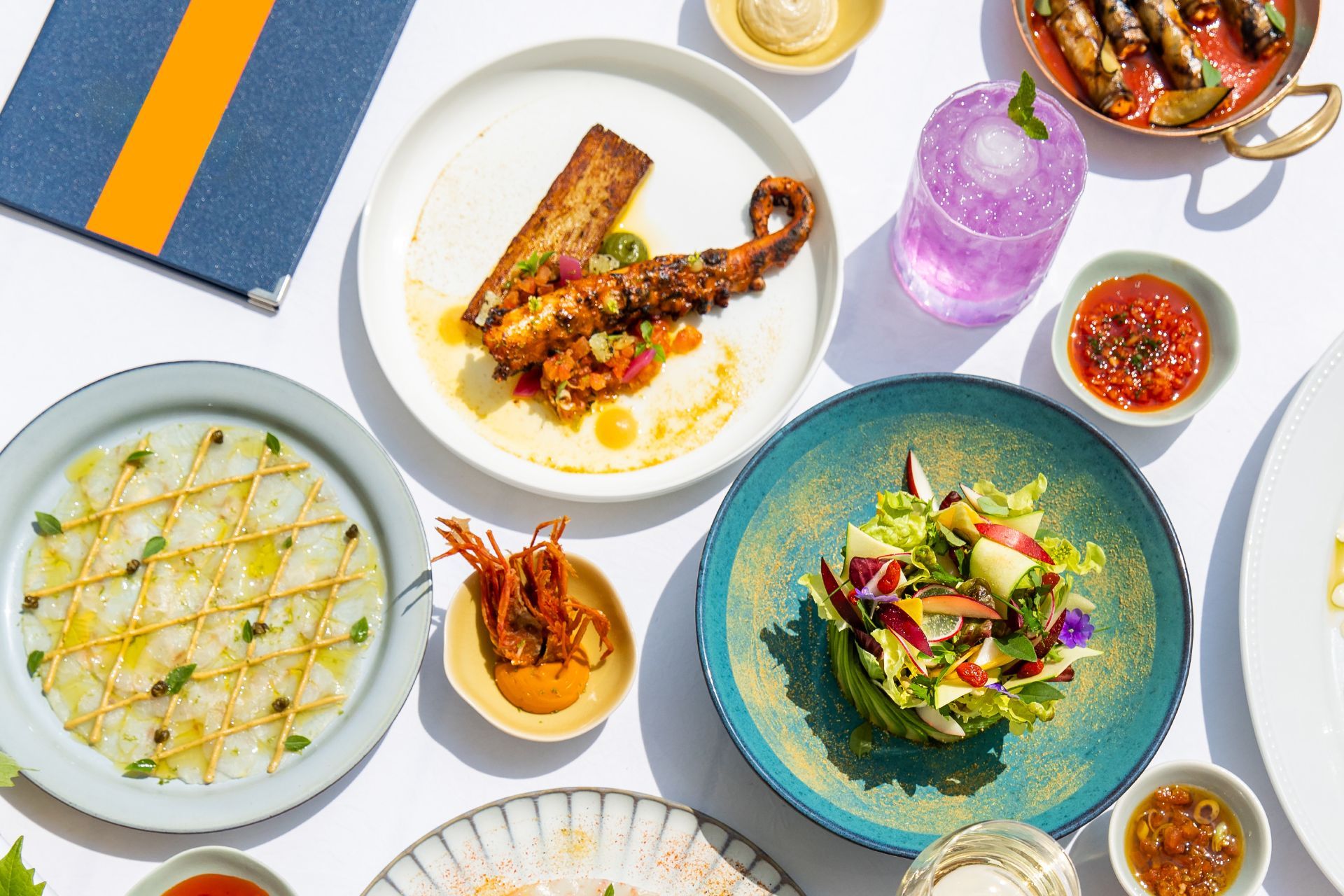 An overhead spread of small plates including grilled octopus, fish carpaccio, a leafy salad, dumplings and a purple cocktail.