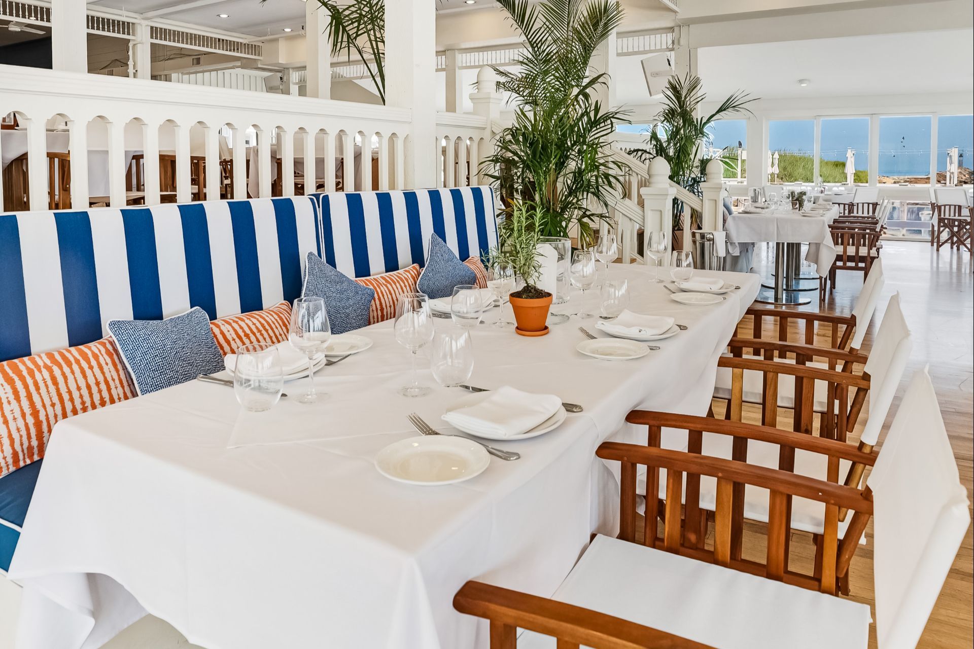 A dining area with a long table set with glassware and striped cushioned benches, overlooking the water.