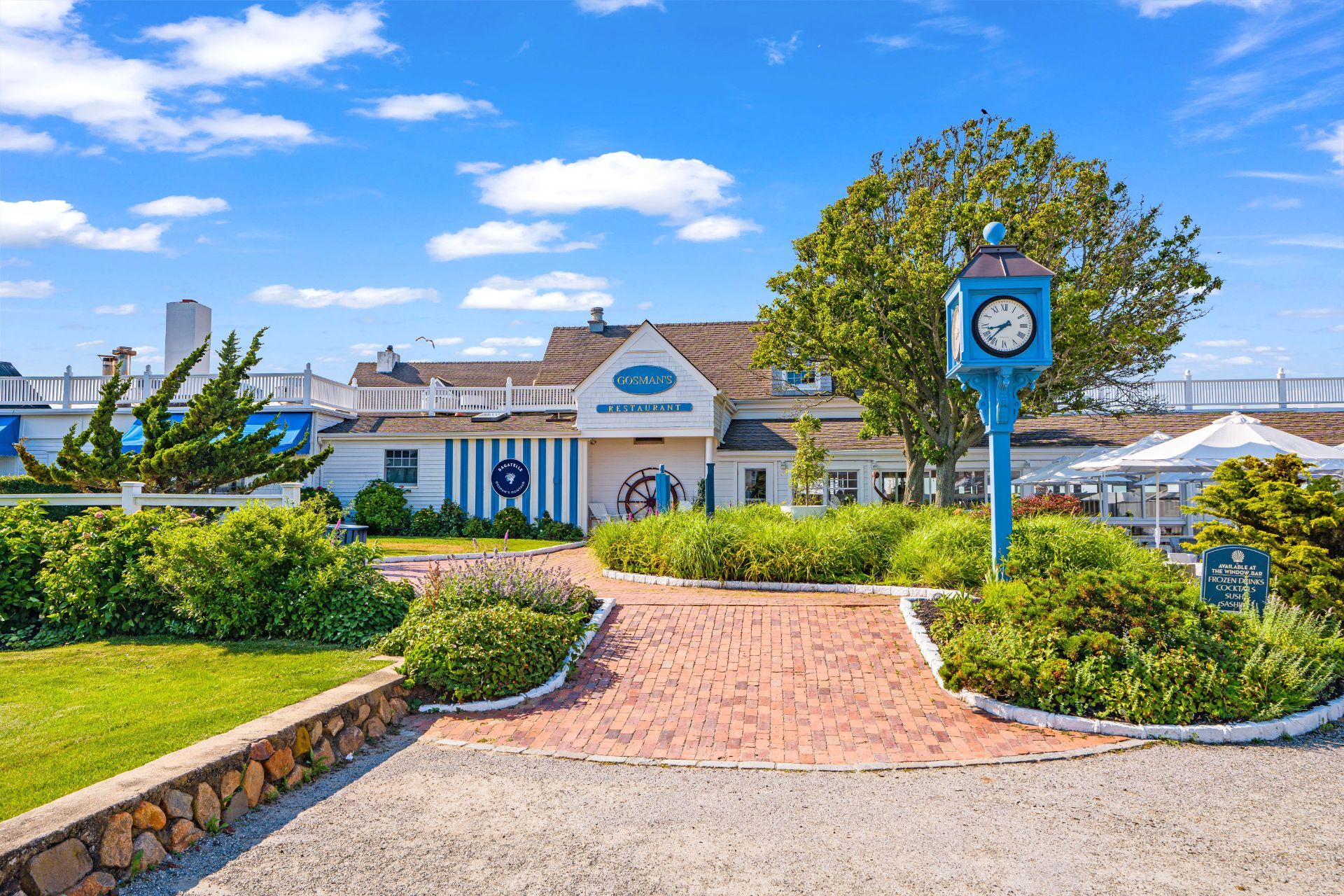 The white and blue exterior of a restaurant with a brick path, landscaped garden and a clock on a post.