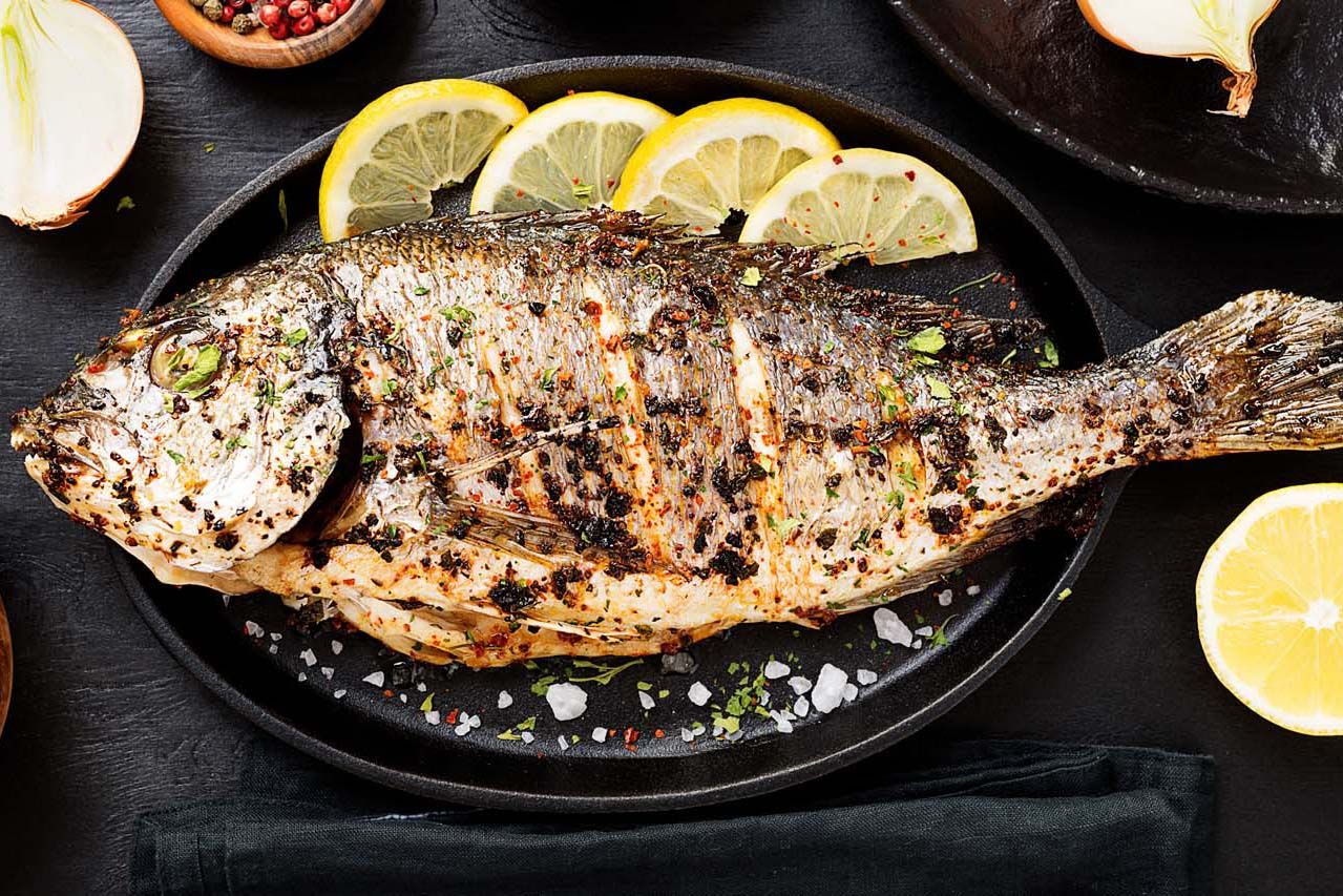 A whole grilled fish seasoned with herbs and spices, served with lemon wedges on a dark plate.