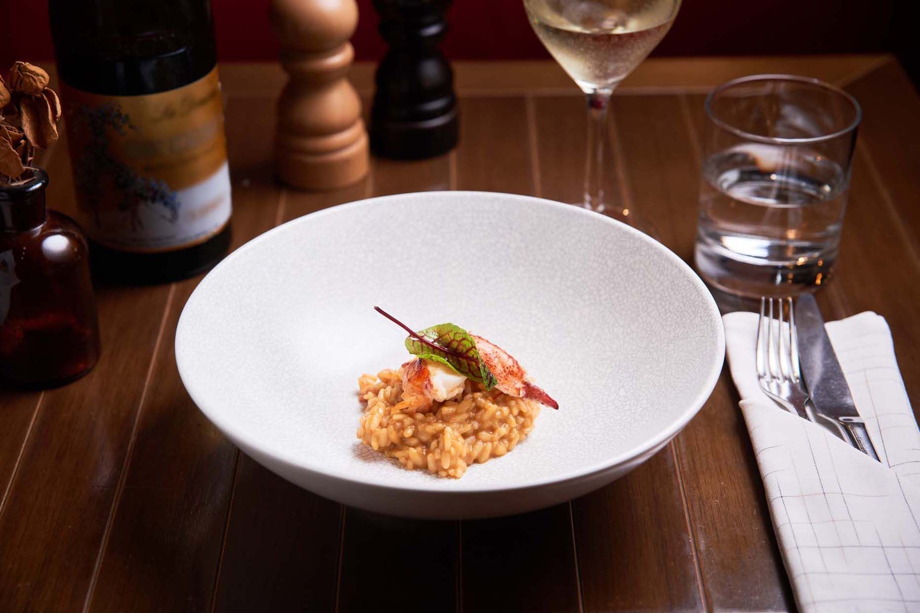 A bowl of risotto topped with seafood and a leaf garnish, set on a table with a wine glass and water glass.