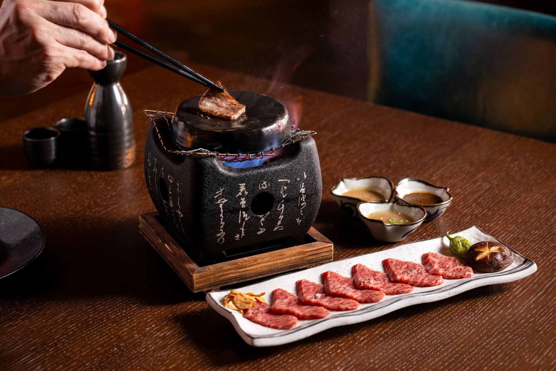 Sliced wagyu beef being seared over a tabletop grill, served alongside raw slices of meat with garlic and a mushroom.