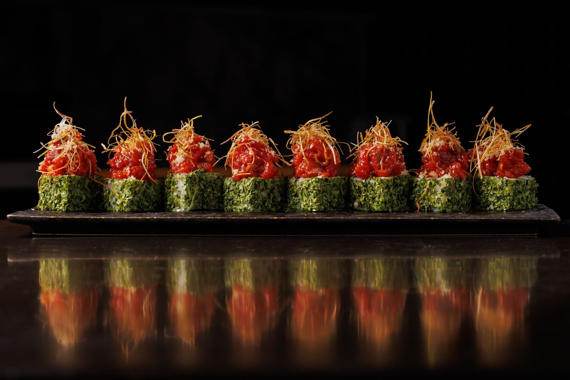 A row of seaweed-wrapped rolls topped with diced tomato and crispy shredded garnish, reflected on a dark surface.
