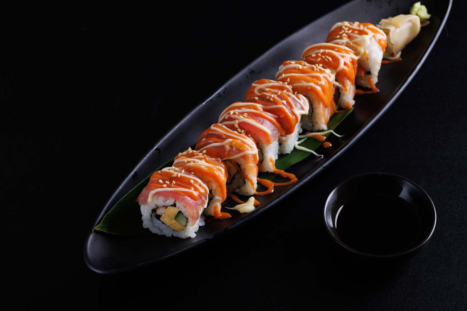 A row of sushi rolls drizzled with sauce and sesame seeds on a boat-shaped plate, served with a small dish of soy sauce.