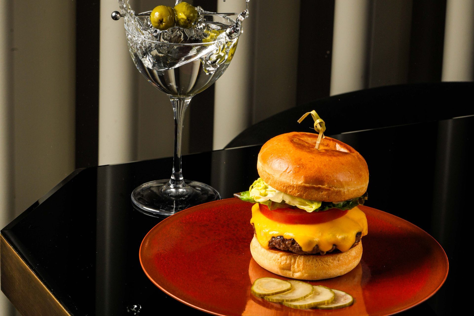 A cheeseburger with melted cheese, tomato, and lettuce on a glazed bun, served with pickles next to a martini glass with olives.