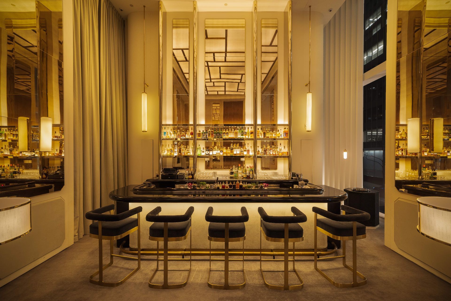 A bar with backlit mirrored shelving, a black counter, and dark upholstered stools with brass legs.