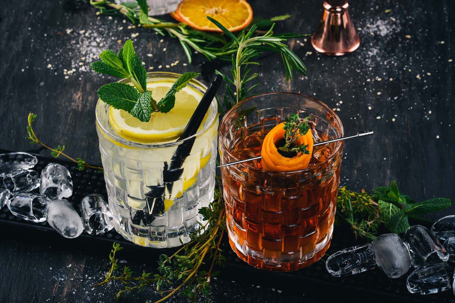Two cocktails on a dark table, one garnished with mint and a lemon slice, the other with a citrus twist and herb sprig, surrounded by ice and fresh herbs.
