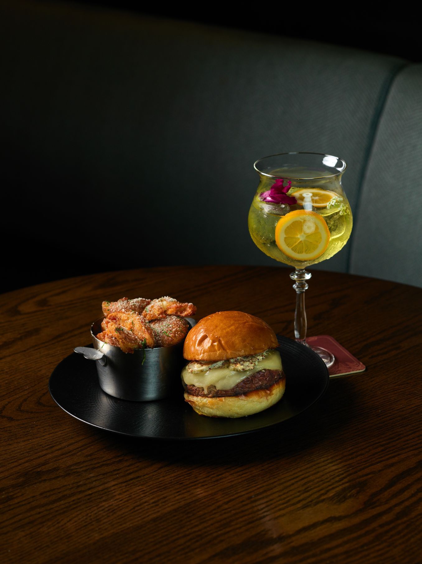 A cheeseburger and a bucket of fried onion rings on a black plate, next to a cocktail garnished with citrus slices and a flower.