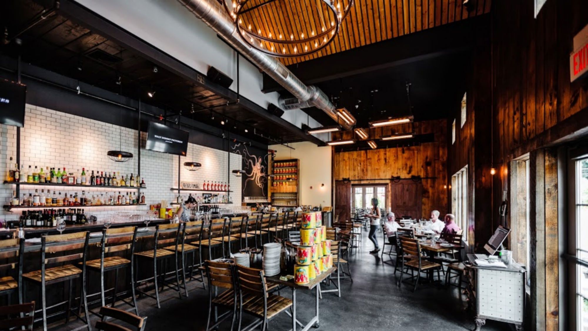 A restaurant with a long bar backed by shelves of bottles, an octopus mural, and a dining area with wood-paneled walls where guests are seated at tables.