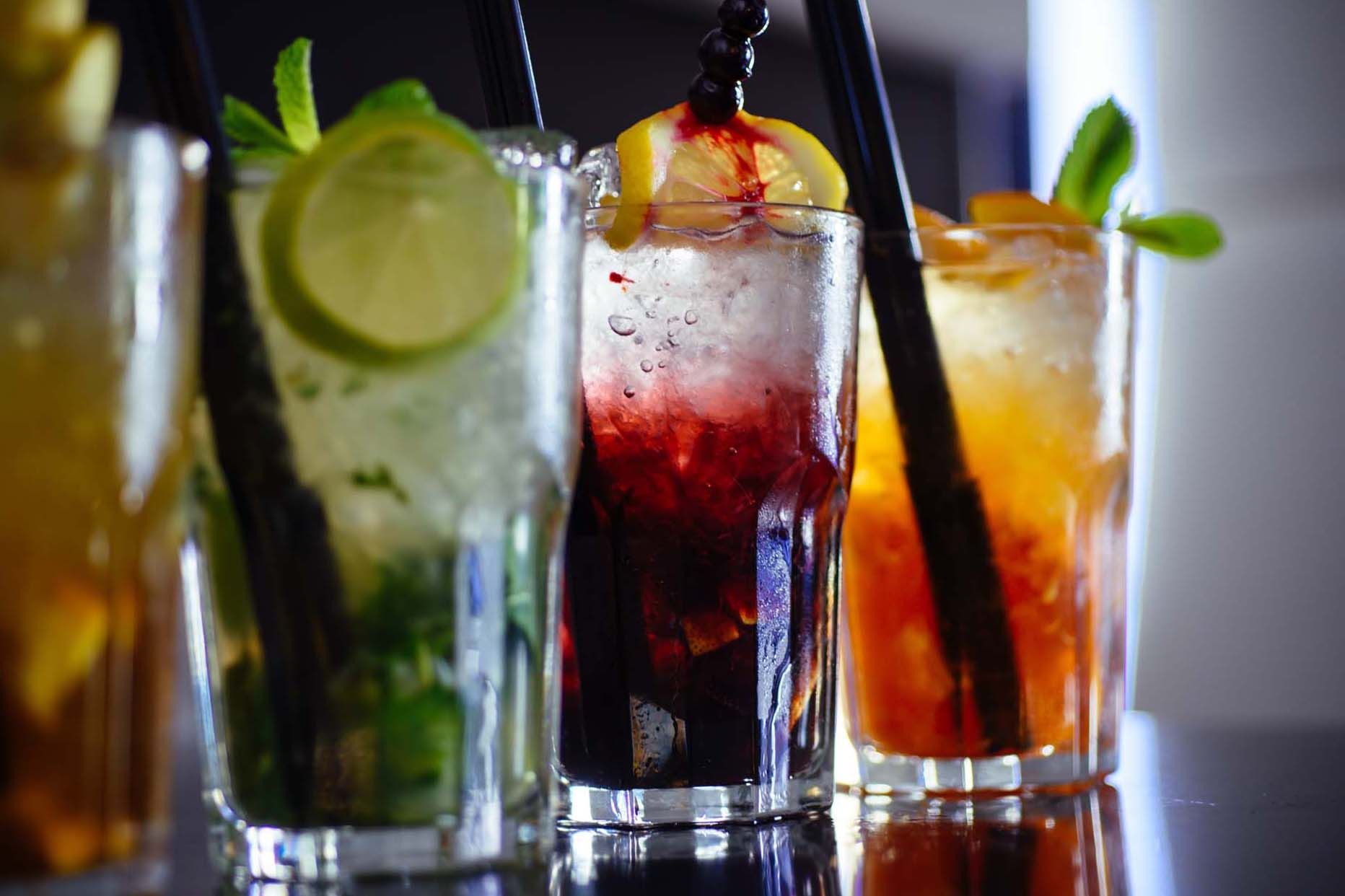 A row of colourful cocktails garnished with citrus slices and mint, served over ice with straws.