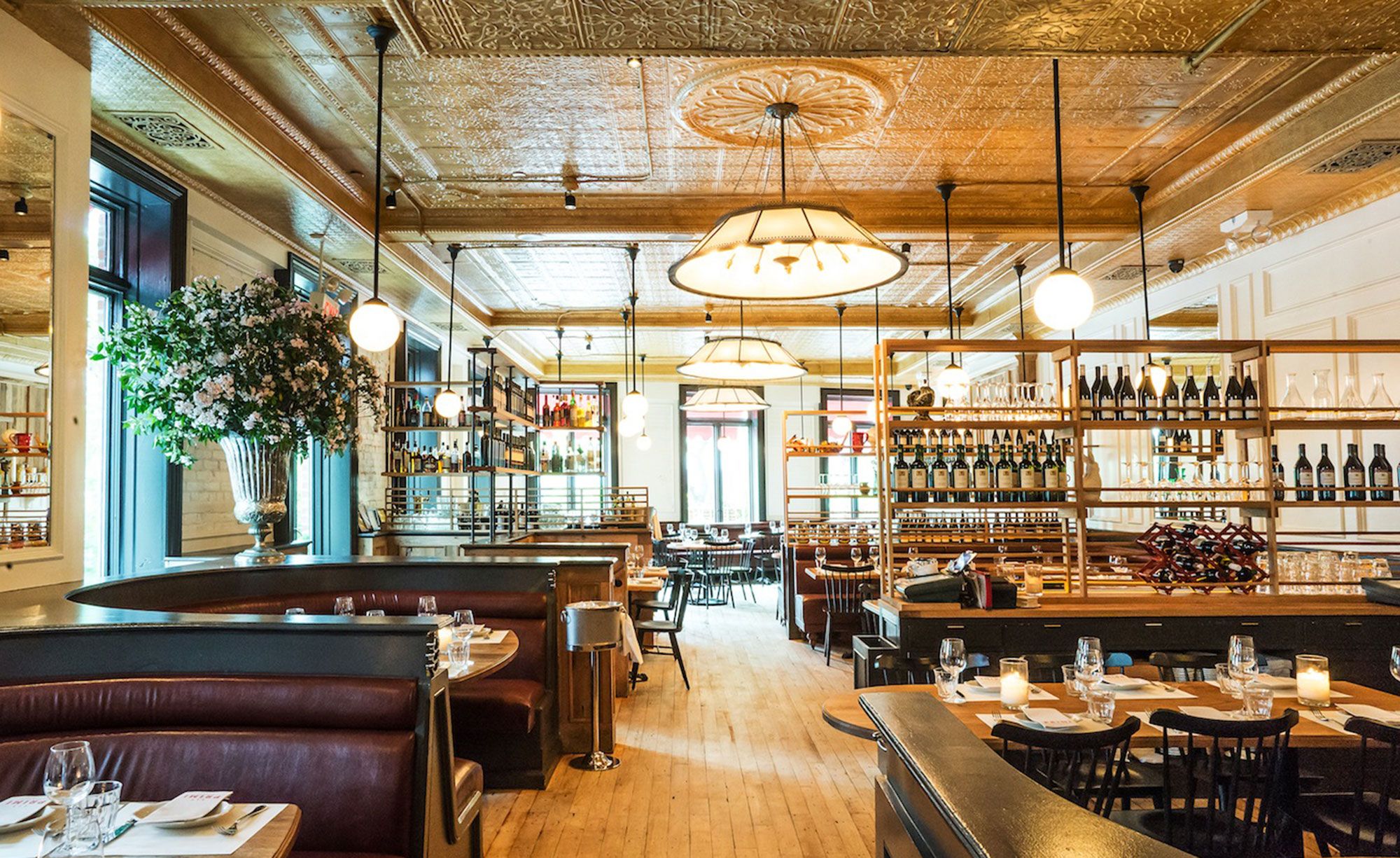 A restaurant dining room with a pressed-tin ceiling, hanging globe lights, and shelves lined with wine bottles.