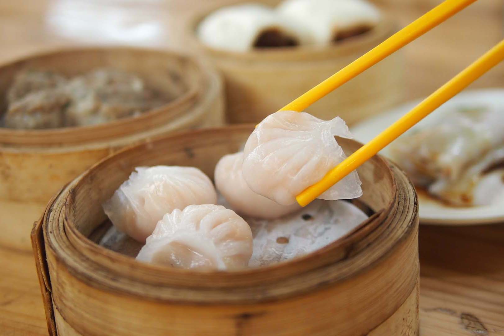Seafood dumplings lifted with chopsticks from a bamboo steamer, with more dim sum in the background.