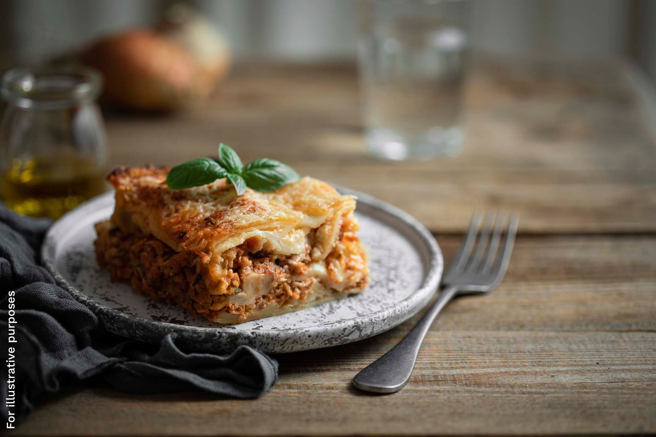 A slice of layered lasagna topped with a basil leaf, served with a fork.