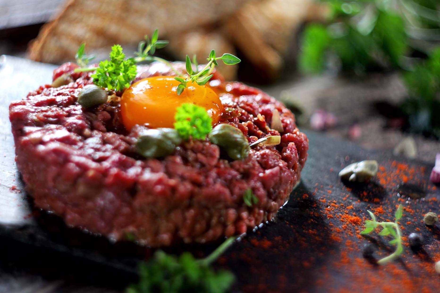 A meat tartare topped with a raw egg yolk, capers and herbs on a dark surface with paprika dusted around it.