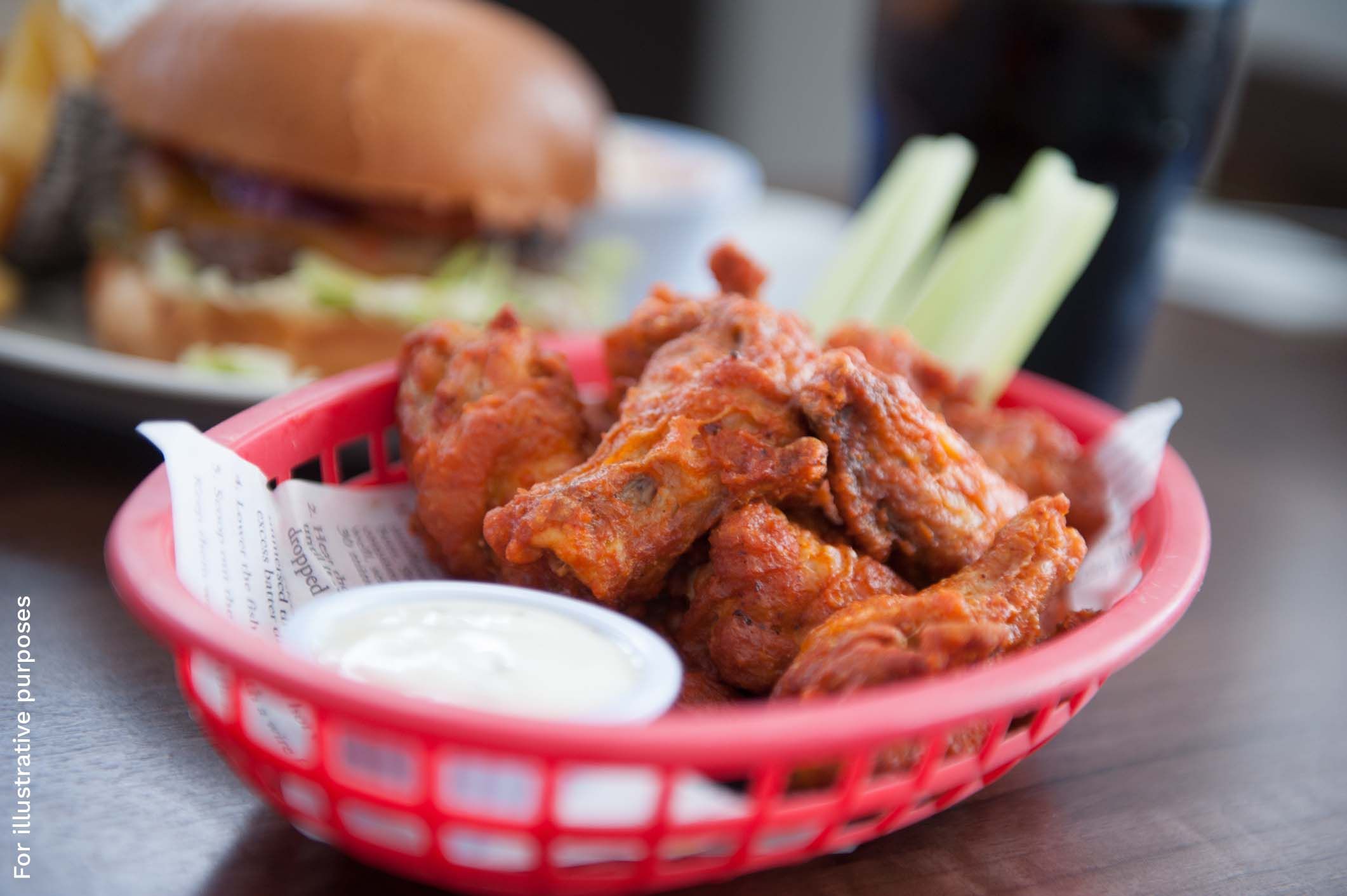 A basket of buffalo wings with celery sticks and a dip, with a burger in the background.