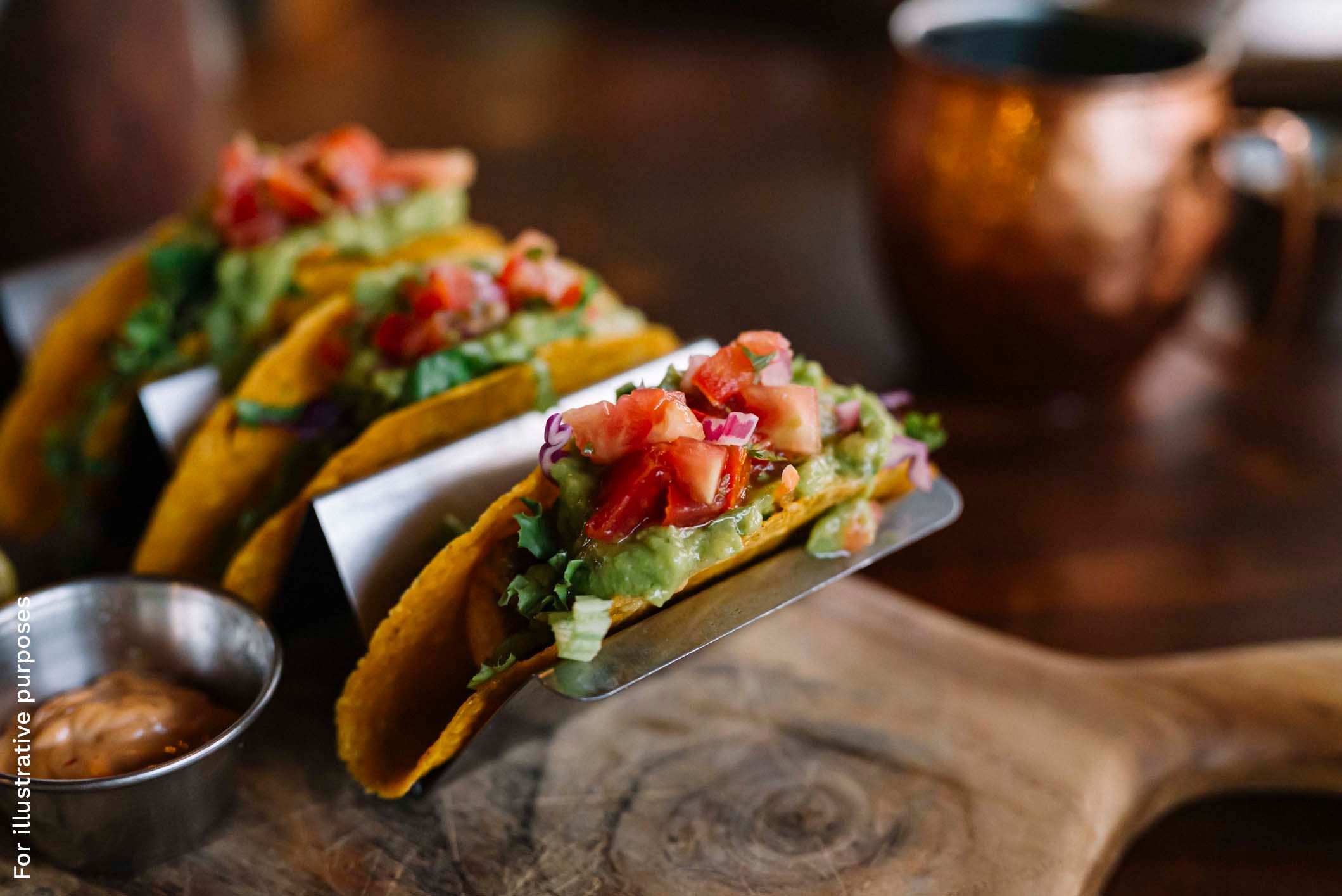 Hard-shell tacos filled with meat, shredded lettuce, guacamole, and diced tomato on a metal stand.